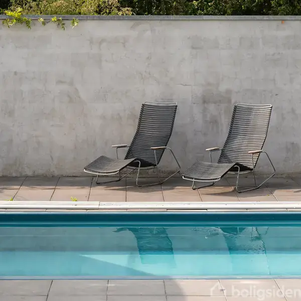 To liggestole står ved siden af hinanden på en flisebelagt terrasse ved en swimmingpool. En mur danner baggrund, og der er lidt grøn beplantning ovenpå muren.