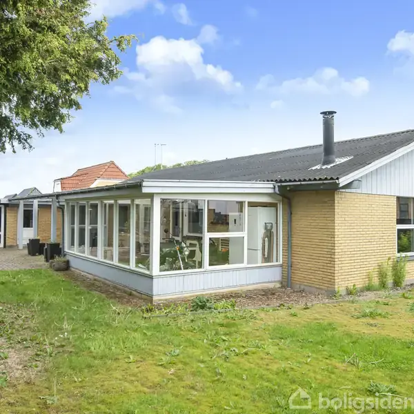 Et enkelt enfamiliehus med gul murstensfacade og udestue mod en grøn have omgivet af buske og træer. Huset har et sort tag og ligger i et roligt boligområde.