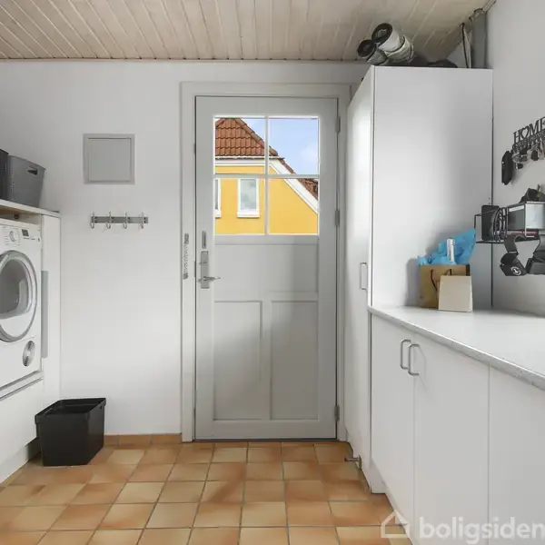En vaskemaskine og tørretumbler står side om side på et vaskerumsgulv af lysebrune fliser. Ved siden af er opbevaringsskabe og en dør med glas. Tekst på væggen: "Home Sweet Home."