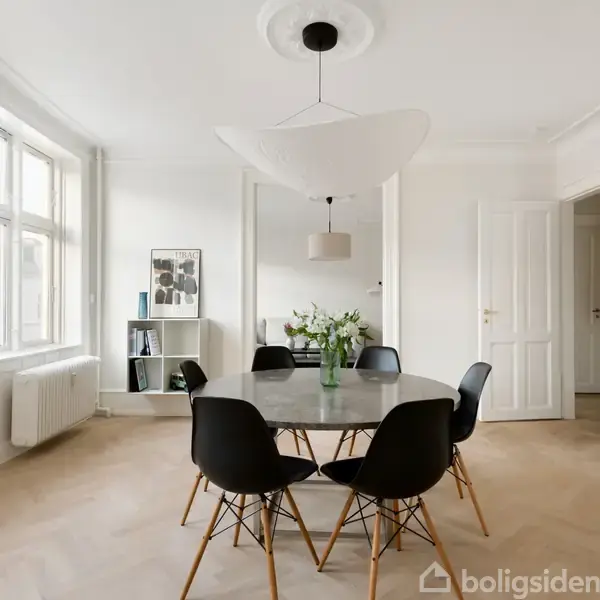 Et rundt bord med blomster i en vase er omgivet af seks sorte stole. Bordet står i en lys spisestue med lyse vægge, trægulv og store vinduer.