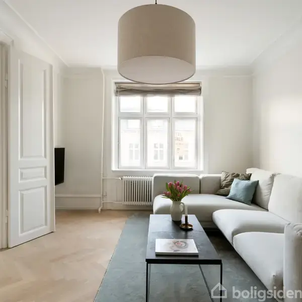 En stor, hvid sofa står i en lys stue ved siden af et sofabord med en vase med blomster. Store vinduer lader lys ind i det minimalistisk indrettede rum.