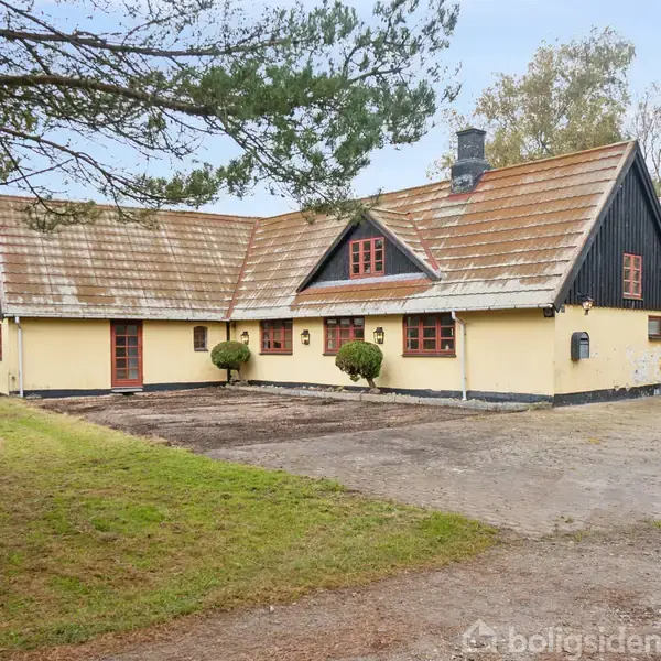 Et gult bindingsværkshus med rødt tag og vinduer står stille i en åbent gårdsplads omgivet af græs og træer i en landlig setting.