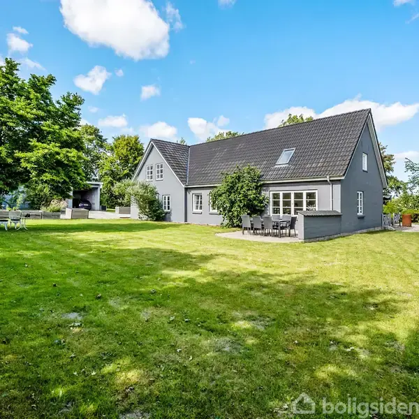 Et gråt, to-etagers hus med sort tag står i en frodig have. Rundt om er der grønne træer og en blå himmel med skyer. En terrasse med havemøbler ses ved huset.