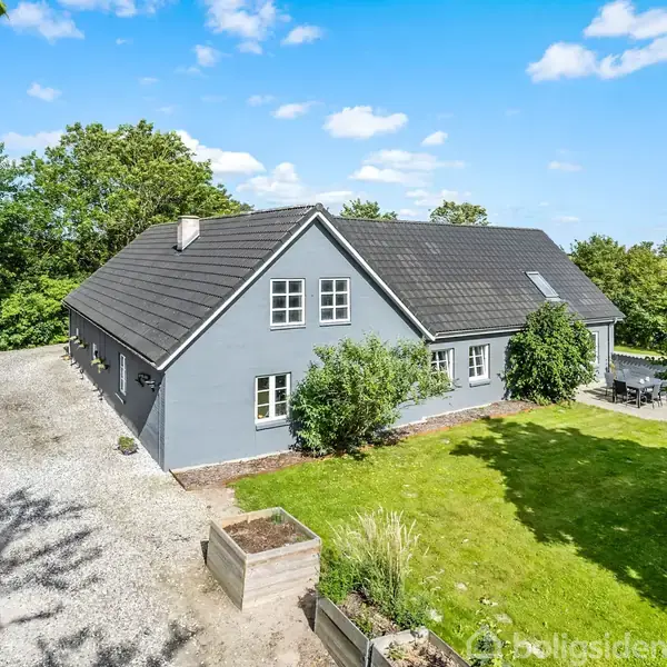 Gråt hus med mørkt tag står midt i en grøn have. Skyfri himmel og træer omkranser ejendommen, med en grusbelagt indkørsel til venstre og terrasse til højre.