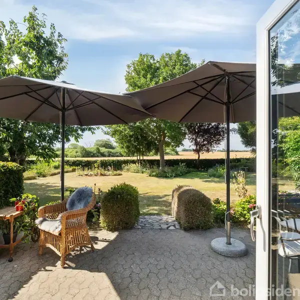 Terrassemøbler står under store parasoller på en flisebelagt terrasse. I baggrunden ses en grøn have med forskellige træer. En åben glasdør leder til terrassen, som er indbydende og lys.