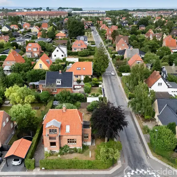 Boliger med røde og sorte tage ligger i et villakvarter. En vej løber gennem området omgivet af træer og haver.