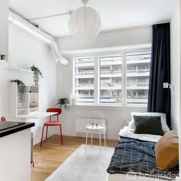 En lys stue indeholder en lille sofa med puder, et rundt bord, og en rød stol ved skrivebordet. Store vinduer lader lys ind, og der hænger en papirlampe i loftet.