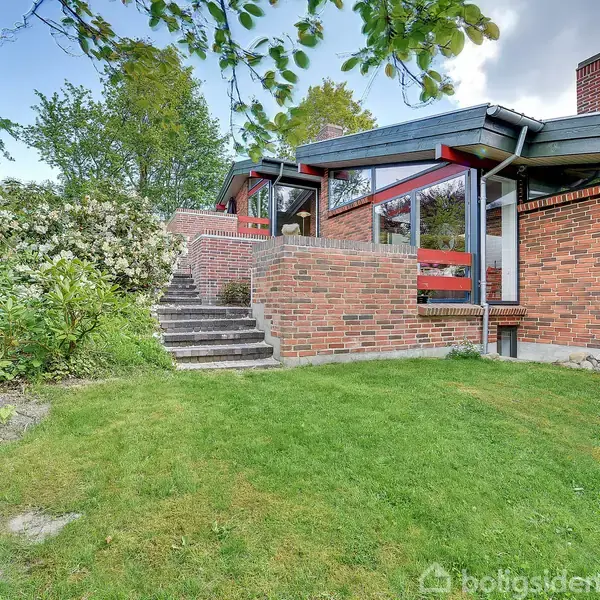 Et rødt murstenshus med store vinduespartier står ved en grøn have. Trapper fører op til huset, omgivet af frodige buske og træer under en blå himmel med enkelte skyer.