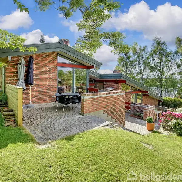 Et moderne hus med røde mursten har en terrasse med bord og stole. Det er omgivet af en frodig have med græsplæne og træer under en blå himmel med skyer.