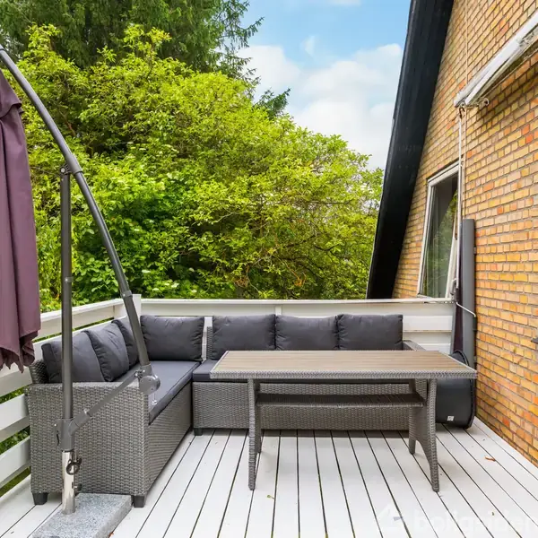 Terrassemøbler med en sort sofa og træbord står stille på en hvid, træbelagt terrasse. En stor parasol er lukket sammen ved siden af. Omkranset af grønne træer og murstensvægge.