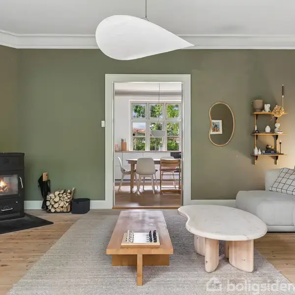 En stue med grå sofa, sofabord og brændeovn. En moderne lampe hænger fra loftet. I baggrunden ses et tilstødende rum med spisebord og stole.