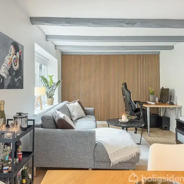 Sofa med puder står foran en væg med træpaneler, ved siden af et skrivebord med stol. Indretningen inkluderer kunst på væggen, en reol med flasker og planter ved vinduet.