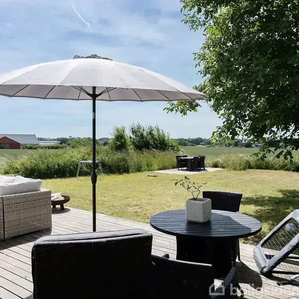 Terrasse møbleret med loungestol, bord med parasol, og grill under åben himmel. Udsigt til græsplæne og marker, omgivet af træer og en basketballkurv.