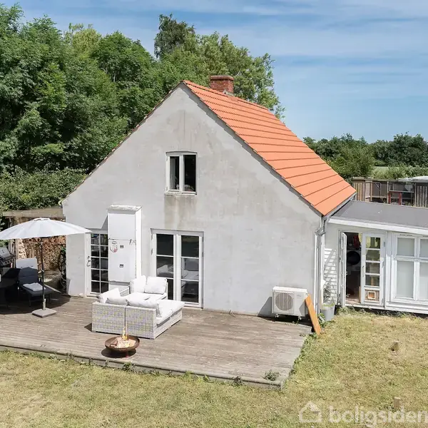 Et hvidt hus med rødt tag står i en have. Huset har en træterrasse med havemøbler og en parasol. Omgivet af træer og en græsplæne.