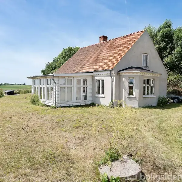 Et charmerende hus med rødt tegltag står stille i en åben, græsset have med få træer og buske i baggrunden under en klar, blå himmel.