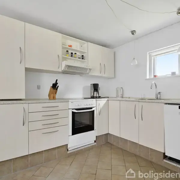 Hvidt køkken indeholder ovn, emhætte, kaffemaskine og knivblok på bordpladen. Fliser på gulvet og lille vindue giver lys. Enkel belysning hænger fra loftet. Tekst "boligsiden" ses ikke.