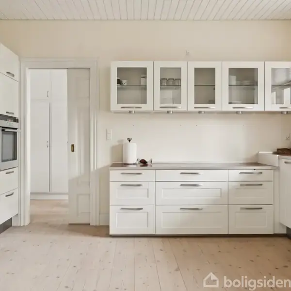 Køkken med hvide skabe, indbygget ovn og skuffer fyldt med køkkenredskaber. I baggrunden er der en dør, der fører til et andet rum. Lys trægulv og hvide vægge.