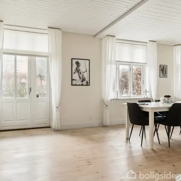 Spisestue med hvidt spisebord og sorte stole står på trægulv. Store vinduer og glasdør med hvide gardiner lader dagslys ind. Væggene er dekoreret med sort-hvide billeder.