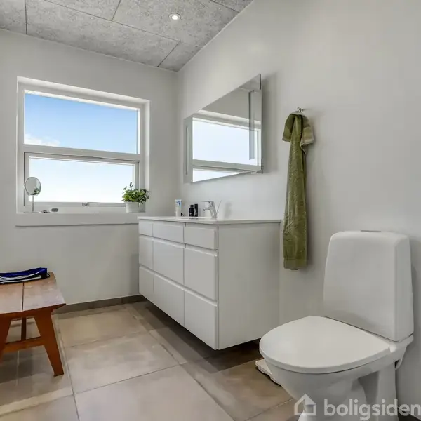 Et lyst og rummeligt badeværelse med toilet og hvidt skab under spejl. En bænk står ved vinduet, og der hænger et håndklæde ved spejlet.