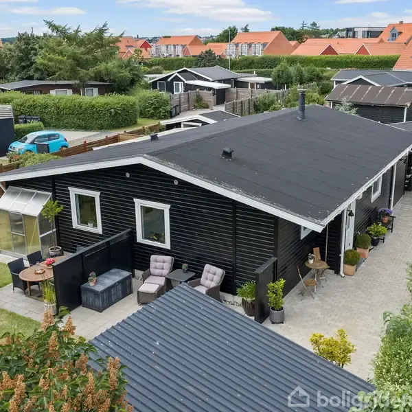 To kurvestole med hynder står ved et sort plankeværk på en flisebelagt terrasse; en blomsterpotte pryder det mellem dem, mens et opbevaringsbænk ses i baggrunden.
