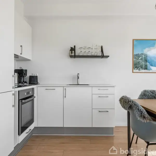 Et moderne køkken med hvide skabe og integreret ovn. Et spisebord med stole med fåreskindsbetræk. Væggen har en hylde med glas og et billede af et bjerglandskab.
