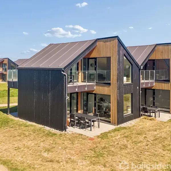 Et moderne sommerhus med sorte træpaneler og store vinduer står i en række af lignende strukturer. Huset har en balkon og en terrasse, og omgivelserne er en græsplæne.