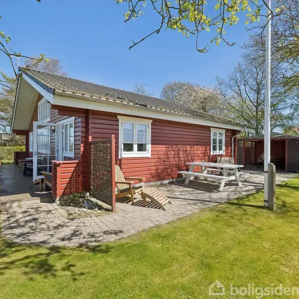 Et rødt træhus med hvide detaljer står på en velplejet græsplæne. Ved siden af er en terrasse med havemøbler og en bænk. Solskin og træer omkranser området.
