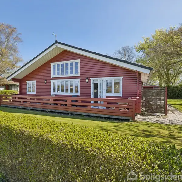 Rødt træhus med store vinduer står i en solrig have. En dansk flagstang er placeret til højre, omgivet af grønne buske og træer.