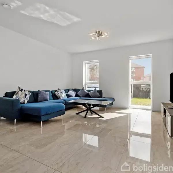 En blå hjørnesofa står i en lys stue med beige flisegulv. Ved siden af sofaen står et sofabord, og en fladskærms-tv er placeret på et lavt skab. To vinduer giver naturligt lys.