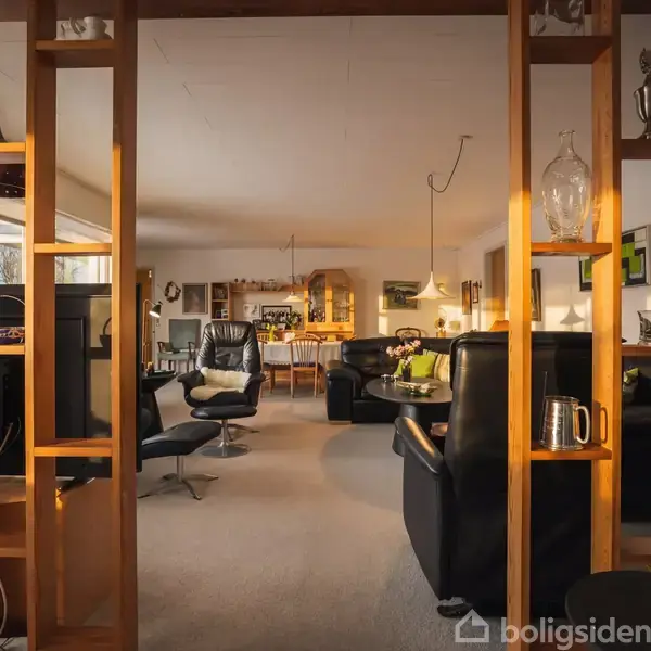 Reol med dekorative genstande opdeler stuearealet; sorte læderstole står i forgrunden, mens spisebordet og flere reoler ses i den lyse og åbne baggrund.