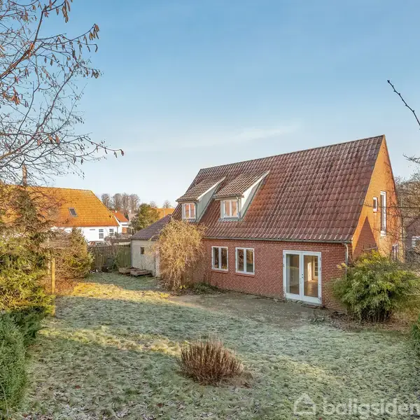 Rødstenshus med skråt tag og hvide vinduer står i en frodig have med græs og buske. Baggrunden har flere træer og huse under en klar, blå himmel.