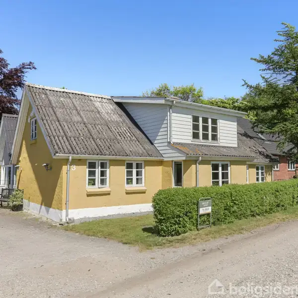 Et gult hus står ved en grusvej, omgivet af en grøn hæk og store træer. Husets tag er gråt, og der ses flere vinduer langs siden. En vejskiltning er placeret ved hækken.