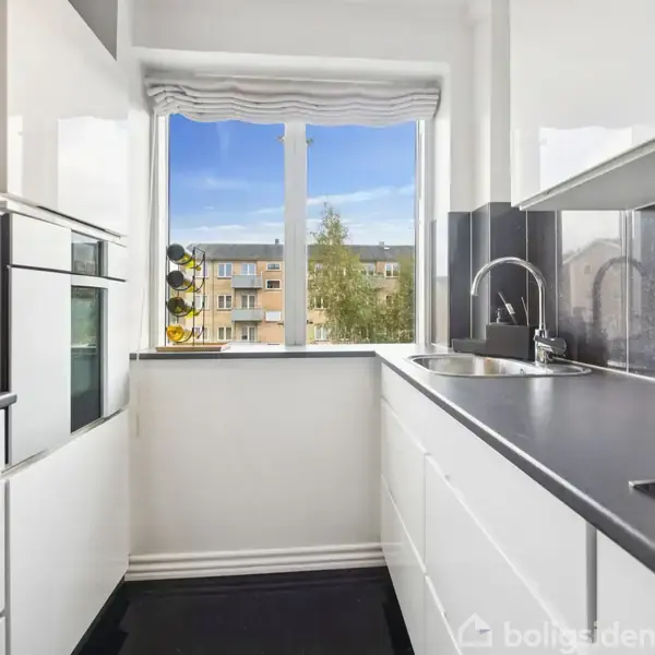 Hvidt, moderne køkken med ovn og vask, set fra en smal vinkel. Et stort vindue med udsigt til bygninger og træer. Flasker står ved vasken, og der er dagslys.