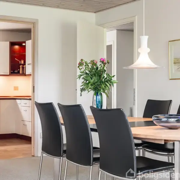 Spisebord med sorte stole omkring sig, placeret i en lys spisestue. Bordet har en vase med blomster. I baggrunden ses en åbning til et køkken med hvide skabe.