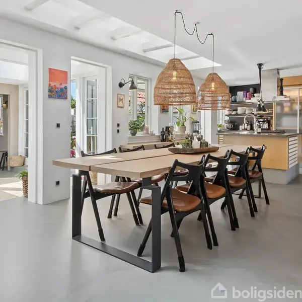 Et langt spisebord med sorte stole står i et lyst, moderne køkken med rå loftsbjælker og store vinduer. To flettede pendellamper hænger over bordet.