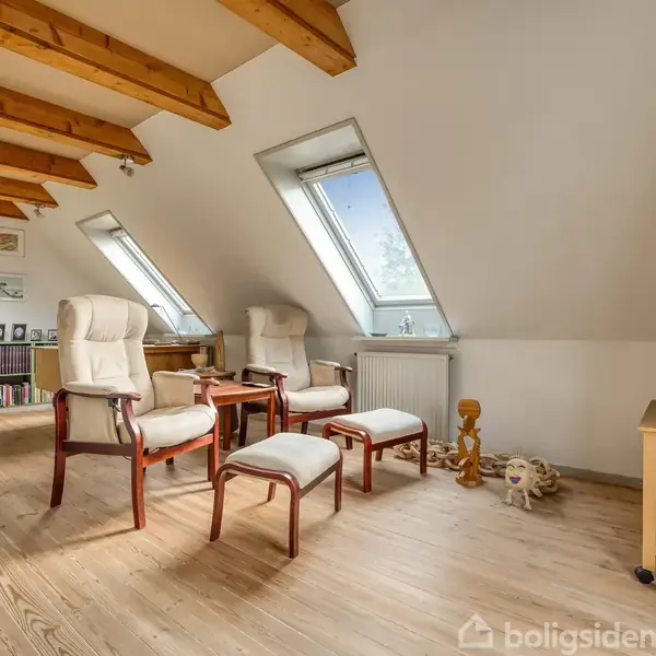 To lænestole og skamler står ved to skråvinduer på et loftsværelse med træbjælker. Et TV står på et lavt bord til højre. En radiator er under vinduerne.