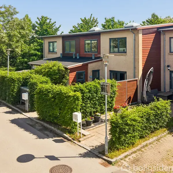 Rækkehus med to etager har en smukt trimmet hæk foran. Huset er omkranset af grønne træer og buske, beliggende i en rolig boligkvarter.