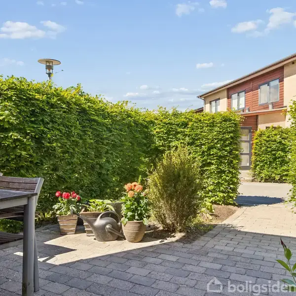 Terrasse med bord og stole i forgrunden, omkranset af grønne buske og blomster i krukker. En bygning med røde mursten og blå himmel i baggrunden.