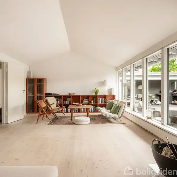 Et rummeligt, lyst stueområde med moderne møbler, herunder stole og sofaer rundt om et sofabord. Store vinduer til venstre giver udsigt til en terrasse med grønt udenfor.