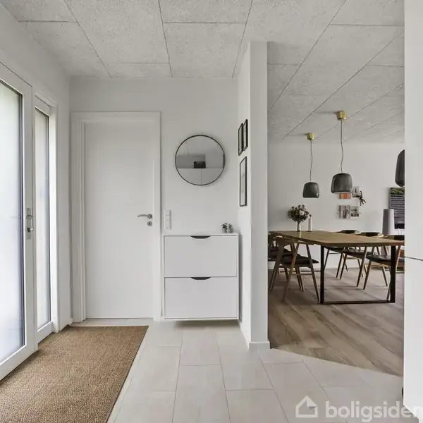En entre viser en hvid skænk og cirkulært spejl, mens dagslys strømmer ind gennem glasdøre. Til højre er en spisestue med træbord, stole og hængelamper, der skaber en moderne atmosfære.
