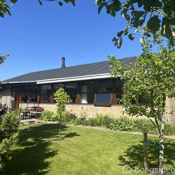 Et én-etagers hus med fladt tag står i en velplejet have. Der er blomsterbede og små træer rundt om. Himlen er klar og blå, hvilket skaber en solrig atmosfære.