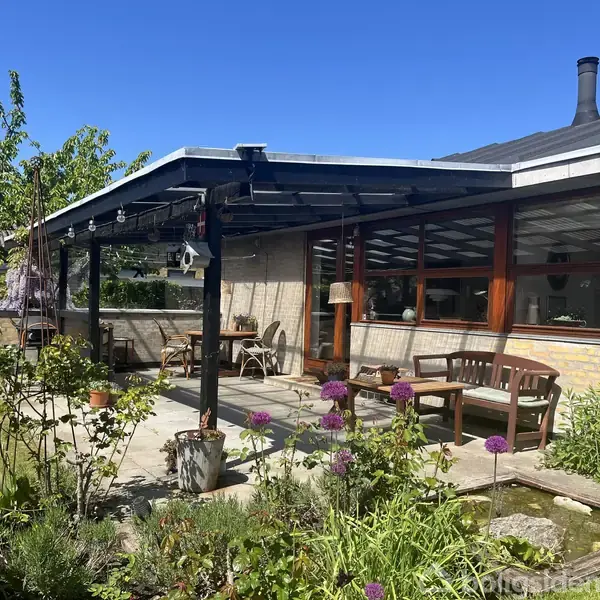 Terrasse under en pergola med træmøbler og planter. Sollys skinner på stenfliserne, mens sprossede vinduer reflekterer lyset. En have med blomster omgiver området, og en lav mur danner grænse.
