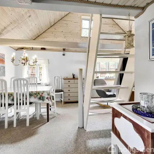 Et hyggeligt rum med spisebord og seks hvide stole står under en loftslampe. I baggrunden ses en stige til hemsen, og væggene er dekoreret med malerier.
