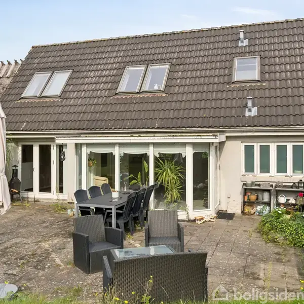 Hus med mørkt tag tækket af tegl, har en terrasse med bord, stole og grill, omgivet af græsplæne og planter.
