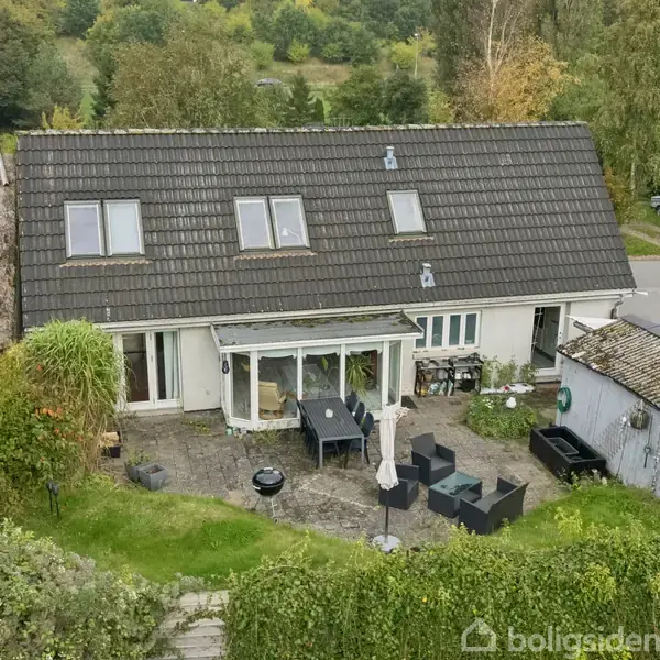 Et hus med et sort tag har en tilhørende terrasse med havemøbler. Terrassen er omgivet af grøn vegetation, beliggende i et skovområde nær en vej.