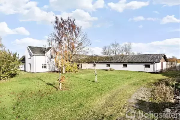 Et hvidt hus står på en grøn græsplæne med spredte træer. Huset har et sort tag, og der er en blå himmel med skyer i baggrunden.