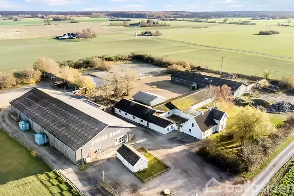 En gård kaldet "TEKAS RANCH" ligger i et åbent landskab med flere bygninger og lader spredt omkring. Omgivet af marker og træer, skaber det en landlig atmosfære.