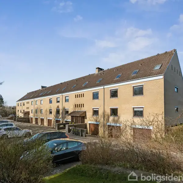 Rækkehus i lysebrun farve med skrå tag og flere vinduer står stille langs en parkering. Omkringliggende buske og en blå himmel udgør omgivelserne.