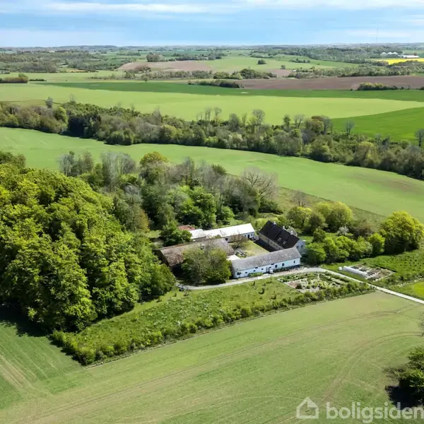 Gårdejendom omkranset af skov og marker. Bygningerne er hvidmalede med mørke tage. Landområdet strækker sig ud i horisonten med grønne og brune marker.