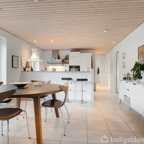 Et moderne køkken-alrum har et rundt spisebord med stole, åbent køkken med højskab og barstole. Rummet er lyst med flisegulv og udsigt til terrassen gennem store vinduer.